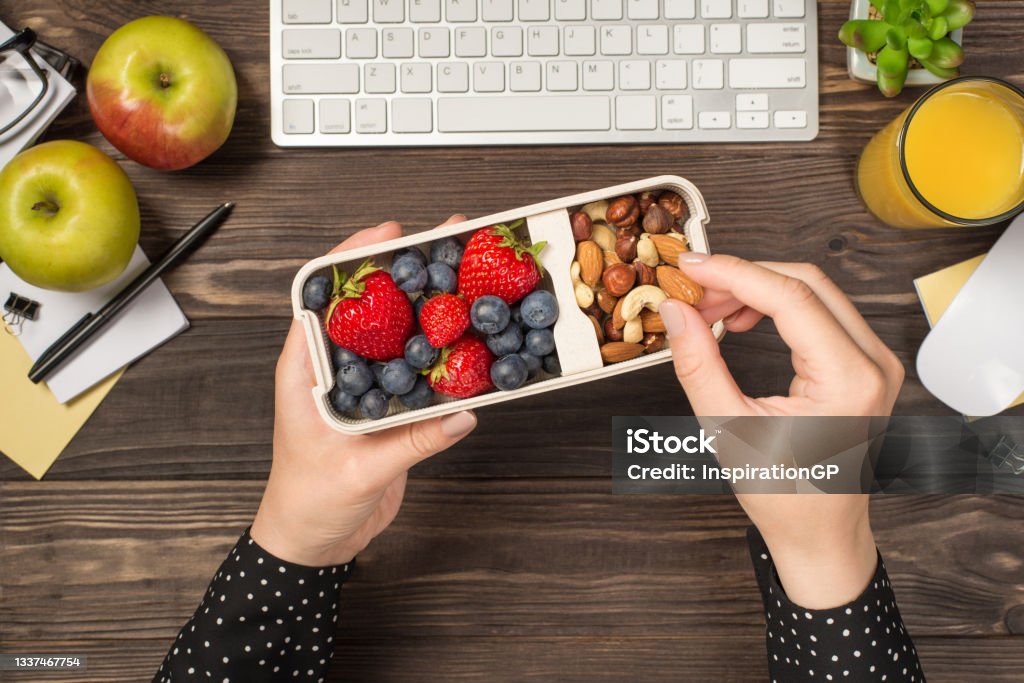 First person top view photo of woman's hands holding lunchbox with healthy meal nuts and berries over apples glass of juice flowerpot stationery keyboard mouse on isolated dark wooden table background